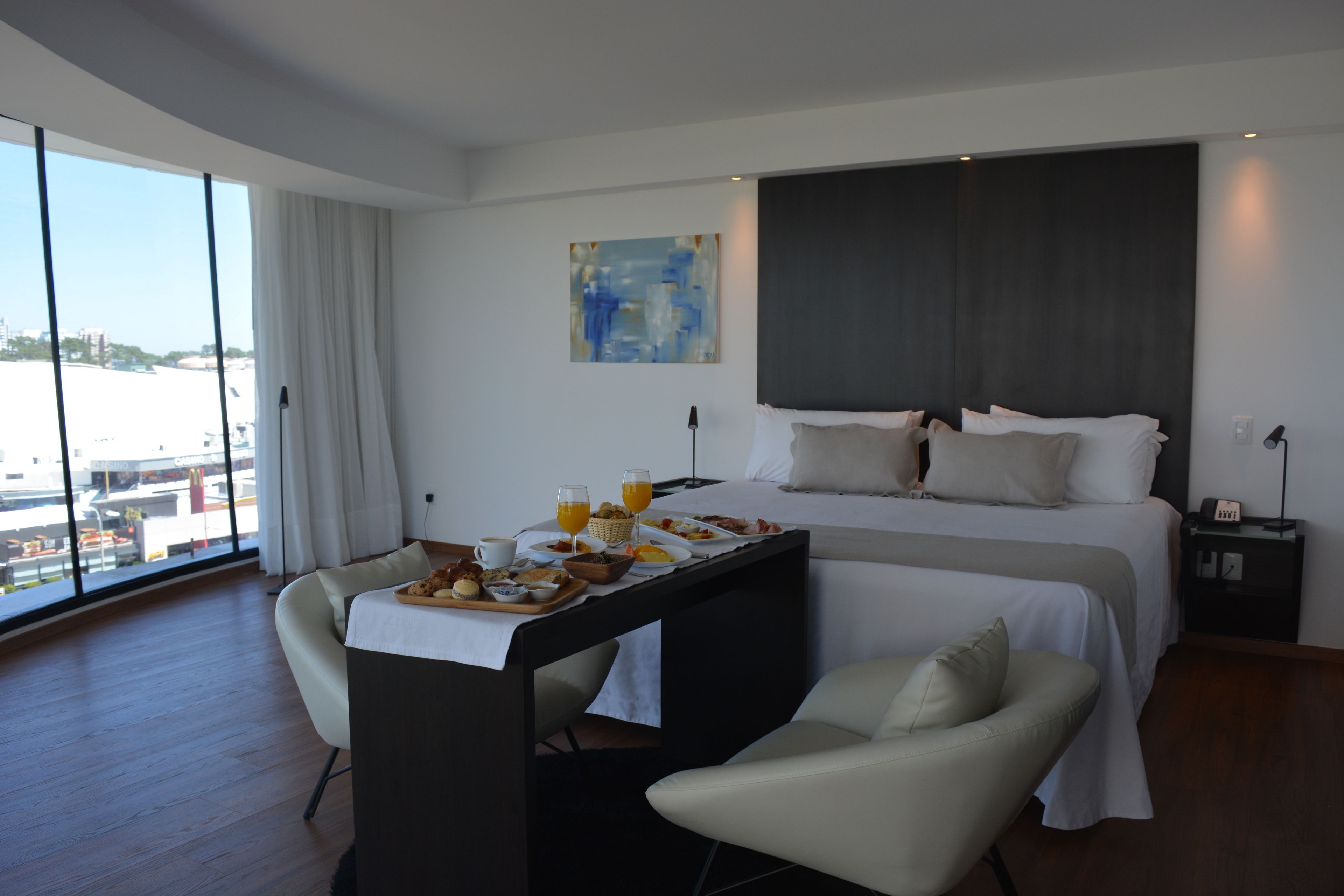 Habitació d'hotel moderna amb un llit gran, una taula de menjador amb esmorzar, vista a la ciutat i un finestral ample.