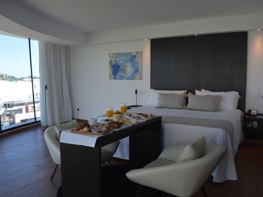 Ein modernes Hotelzimmer mit einem großen Bett, einem Esstisch mit Frühstück, Blick auf die Stadt und einem großen Fenster.