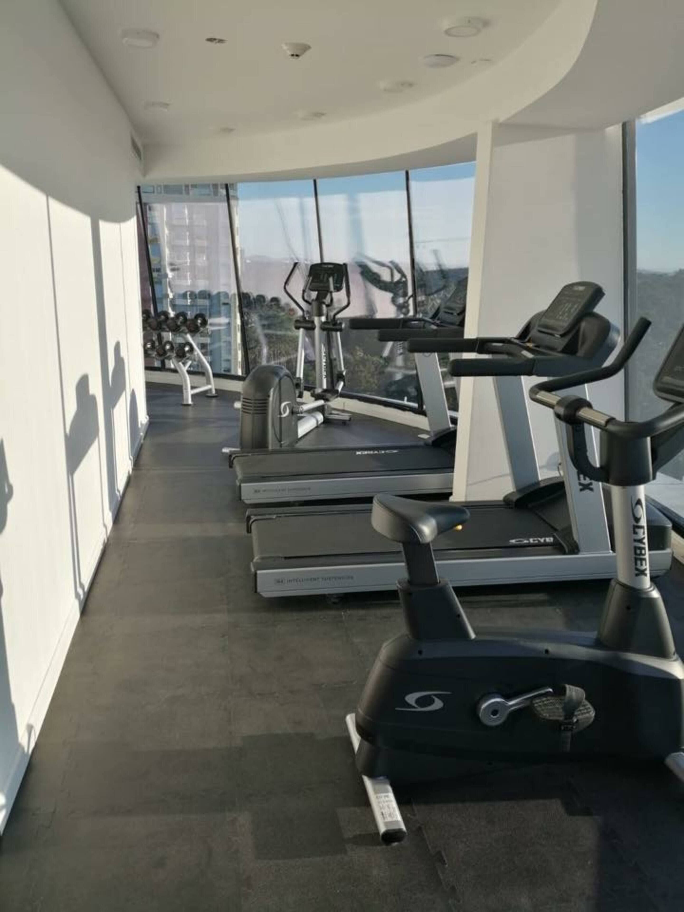 Une salle de sport équipée de tapis de course et de vélos elliptiques, dotée de grandes fenêtres et d’un plafond haut.
