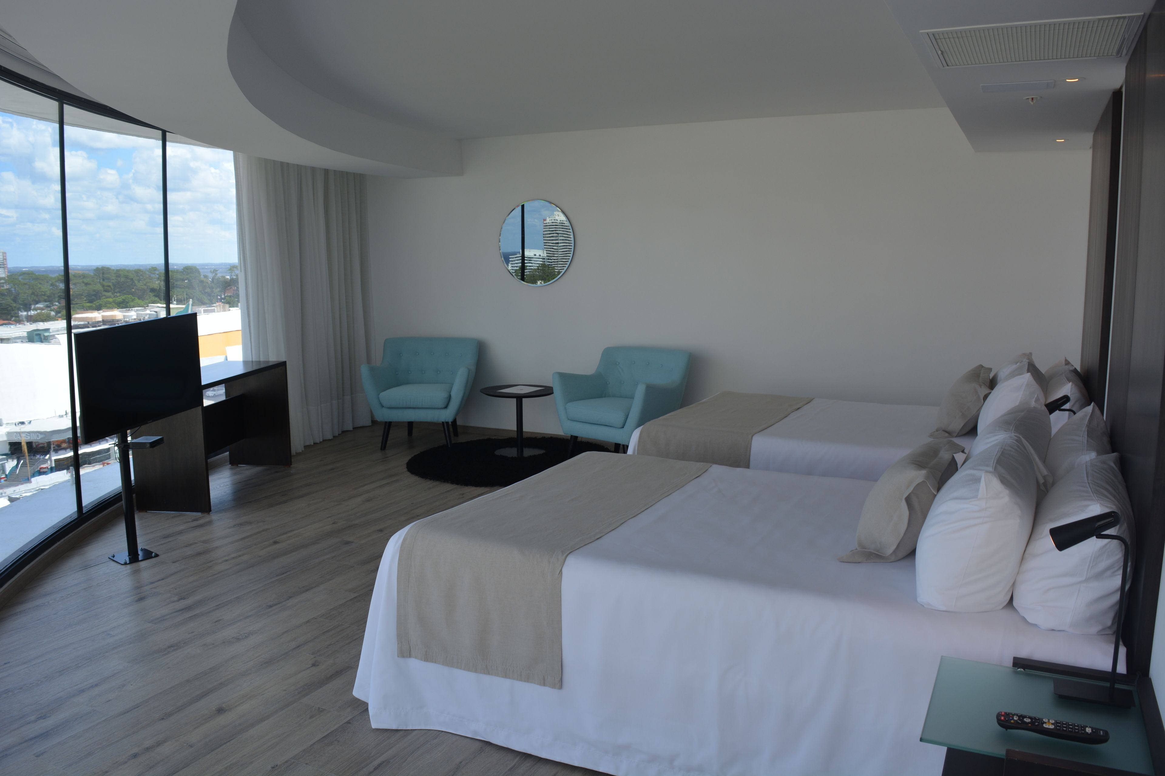 Habitación de hotel moderna con una cama grande, dos sillones, una mesita, un televisor y vistas a la ciudad.