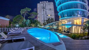 A swimming pool area with lounge chairs and a modern building with a curved glass facade. - Don Majestic Hotel Punta del Este (Punta del Este)