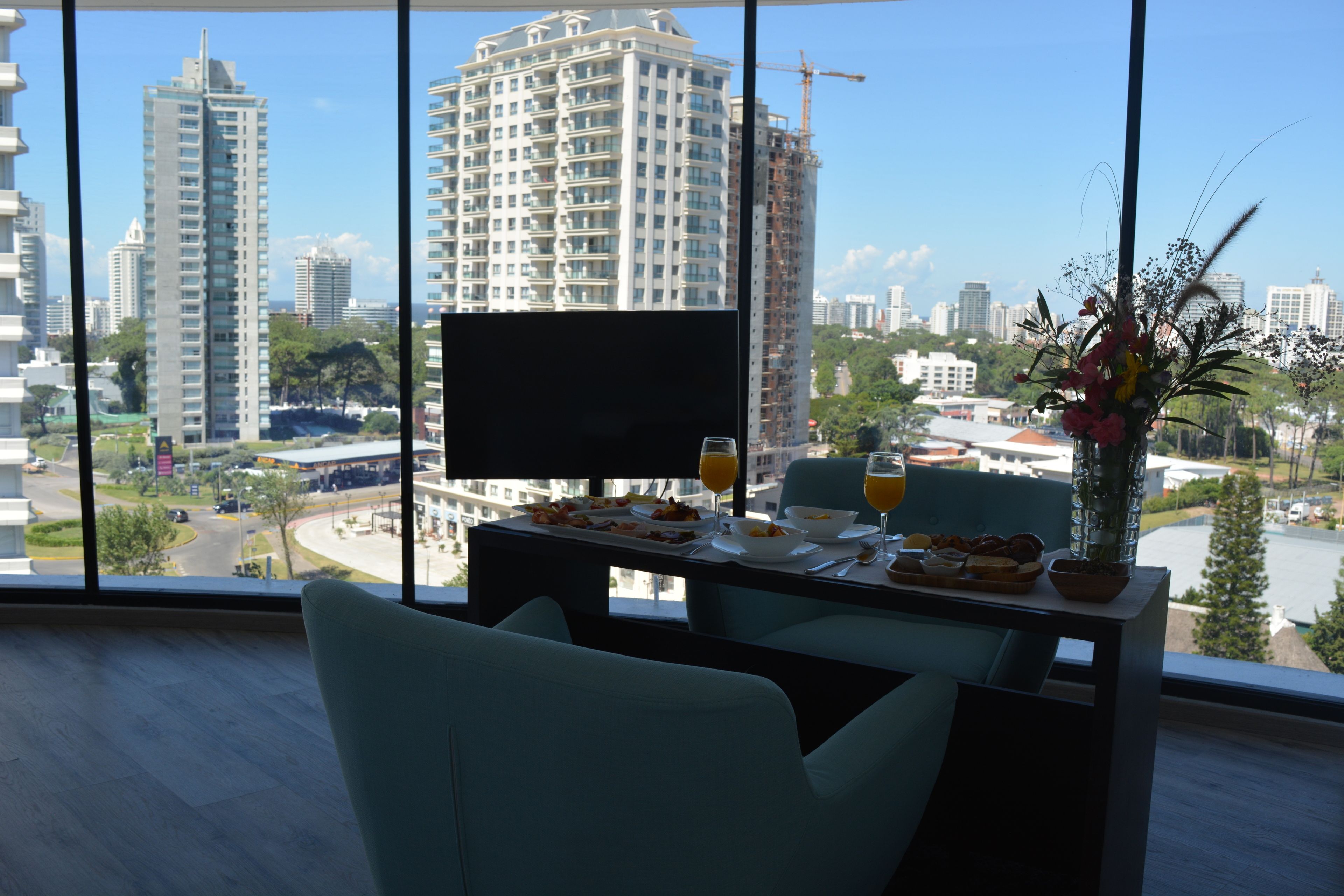 Un comedor con una mesa preparada para dos personas, un televisor de pantalla plana y vistas de la ciudad que incluyen edificios altos y una obra en construcción.