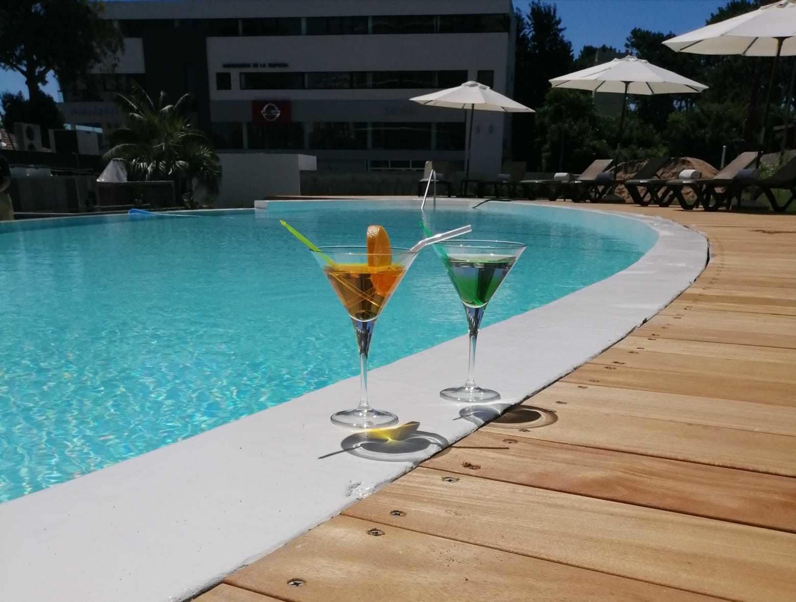 Zona junto a la piscina con dos vasos de martini, uno con una bebida amarilla y el otro con una verde, y una terraza de madera.