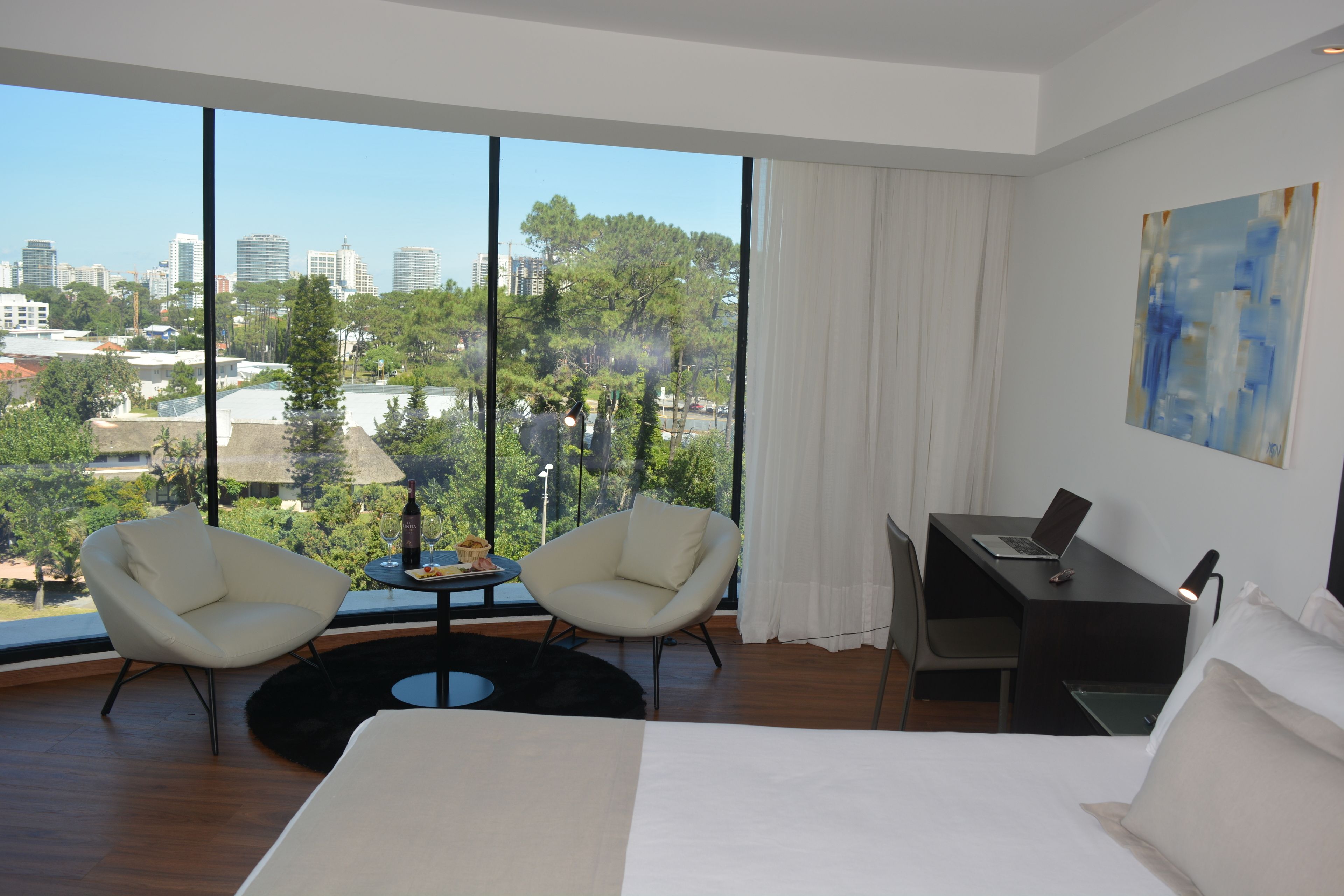 Une chambre d’hôtel moderne dotée d’un grand lit, de deux fauteuils, d’un bureau avec un ordinateur portable et offrant une vue sur la ville grâce à de grandes fenêtres.