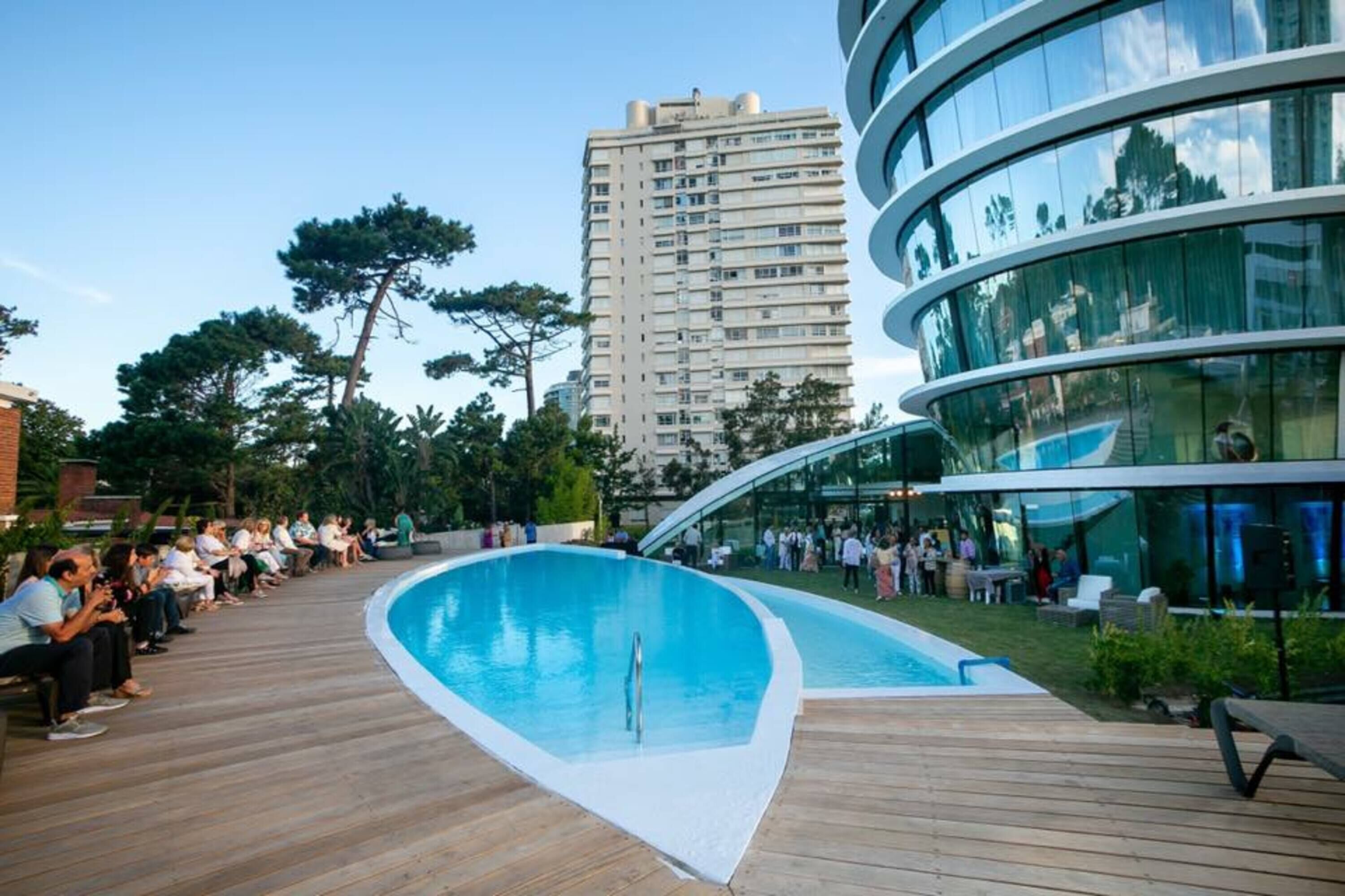 Een modern zwembadgedeelte met een houten terras, omgeven door een glazen gebouw en een hoog woongebouw op de achtergrond.