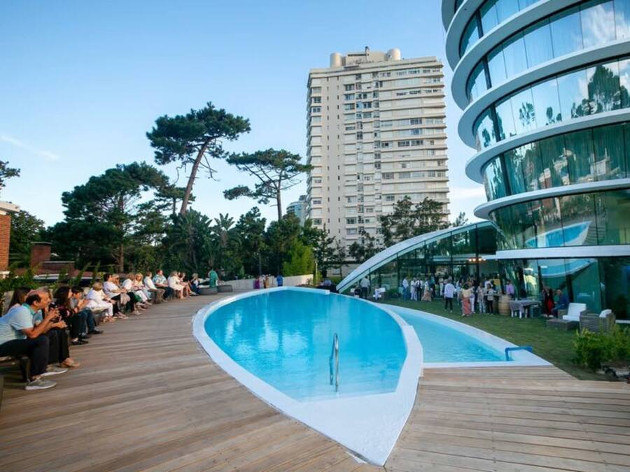 Un área de piscina moderna con cubierta de madera, rodeada por un edificio de cristal y un edificio residencial alto al fondo.