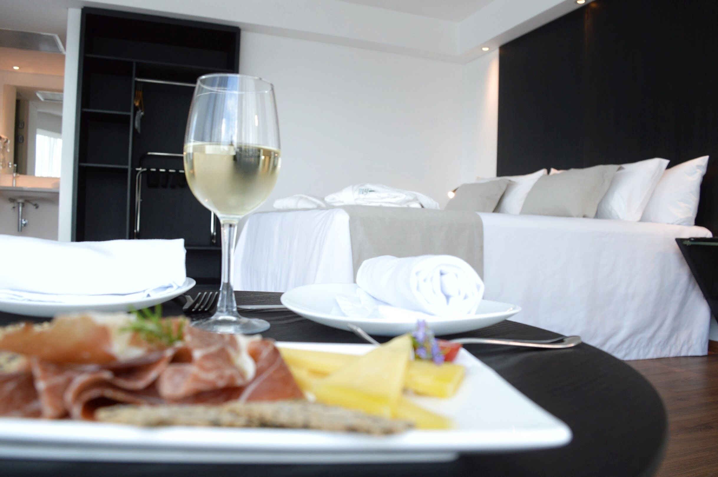 A hotel room with a neatly made bed, a dining table with a plate of food and a glass of white wine, and a black wall.