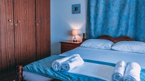 A hotel room with a wooden wardrobe, a bed with blue bedding, a nightstand with a lamp, and a blue curtain. - Casa Di Bella (Volos)