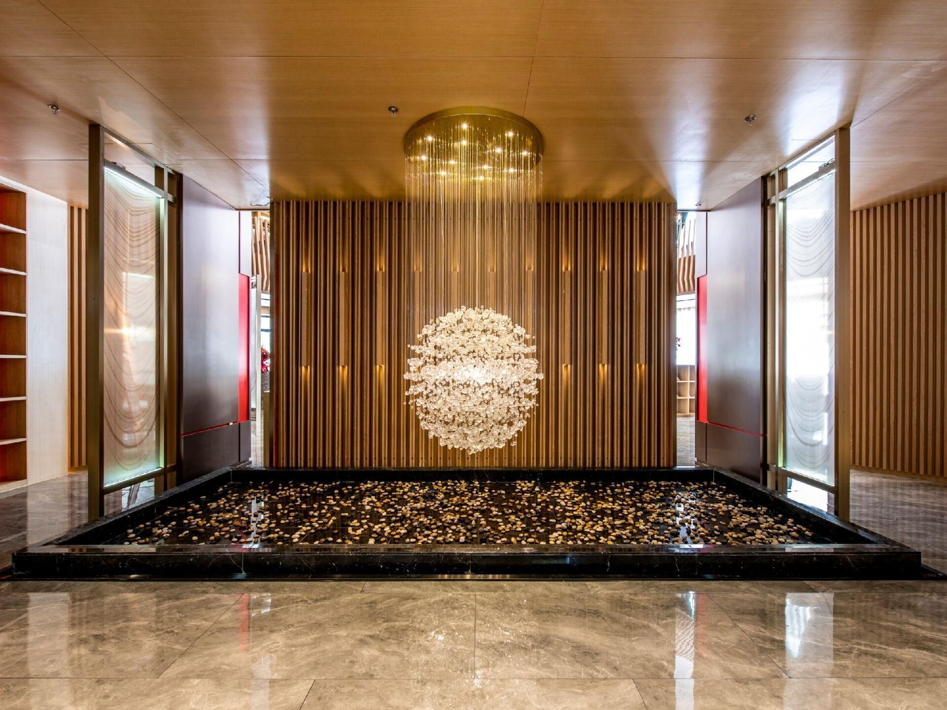 A spacious lobby with a reflective marble floor, a central water feature with floating pebbles, and a modern chandelier.