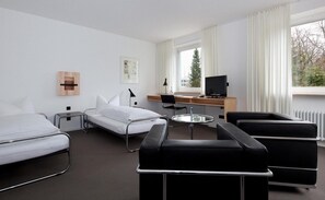 A hotel room with two beds, a desk with a computer, a chair, and a view of trees through the window. - Akademie Der Dioezese (Stuttgart)