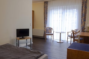 A hotel room with a bed, a TV on a stand, a small table, two chairs, a round table, and a window with curtains.