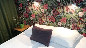 A bed with white linens and a brown pillow, a green plant on a wooden nightstand, and a floral wallpaper.