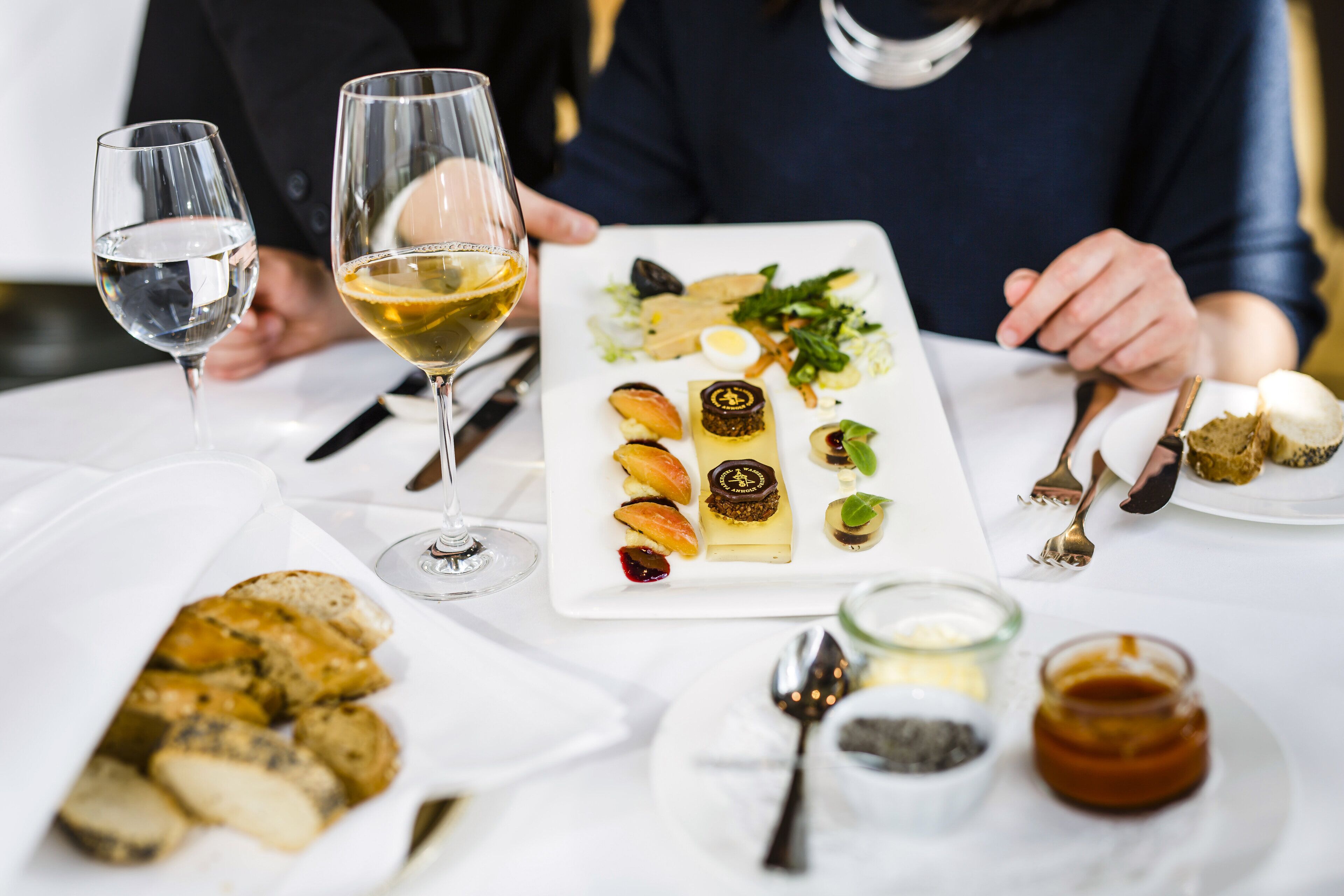 A dining table with a variety of food items and drinks, including a white plate with assorted appetizers, a glass of white wine, and a glass of water.