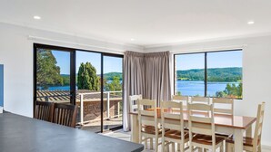 Un coin repas avec une table et des chaises, de grandes fenêtres donnant sur un lac et des arbres, et un balcon avec une balustrade.