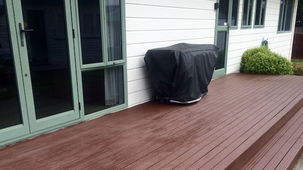 Una terraza de madera con una zona de parilla cubierta, un ventanal corredero y un pequeño arbusto.