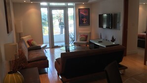 A modern living room with a sofa, armchair, and a glass table. There is a TV mounted on the wall and a door leading to a balcony. - Miami Beach Luxury on South Beach Suite 303 (Miami Beach)