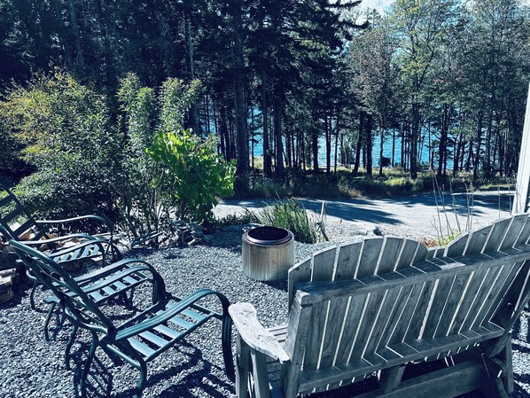 A seating area with Adirondack chairs and a fire pit, surrounded by dense forest and a view of a lake. - Private Ocean Beach Front House (Hancock)