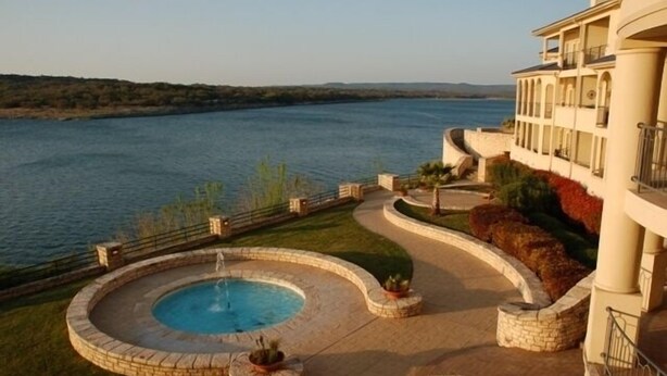A pool with a fountain, surrounded by a landscaped area with a view of a body of water and hills in the background. - Luxury Condo on its Own Private Island on Lake Travis (Lago Vista)