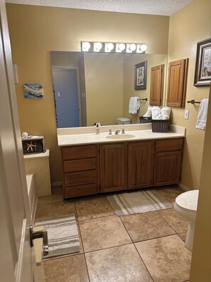 A bathroom with a double sink vanity, wooden cabinets, a large mirror with lights, a toilet, and a rug.