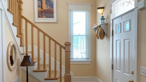 A wooden staircase with a handrail, a framed picture on the wall, a lamp on a table, and a white door with a decorative handle. - OCEAN BLOCK IN SOUTH REHOBOTH! (REHOBOTH BEACH)