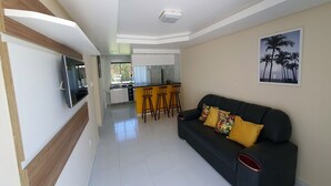 Una sala moderna con un sofá negro, un televisor montado en la pared y una cocina al fondo.