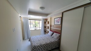 Un dormitorio con cama, armario, ventana con cortinas y estante con objetos decorativos.