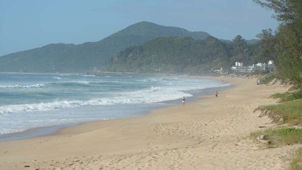 Ein Strand mit einem Berg im Hintergrund, sandigem Ufer und einigen Spaziergängern.