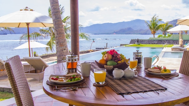 A wooden table with breakfast items, a view of the ocean, and lounge chairs with umbrellas.