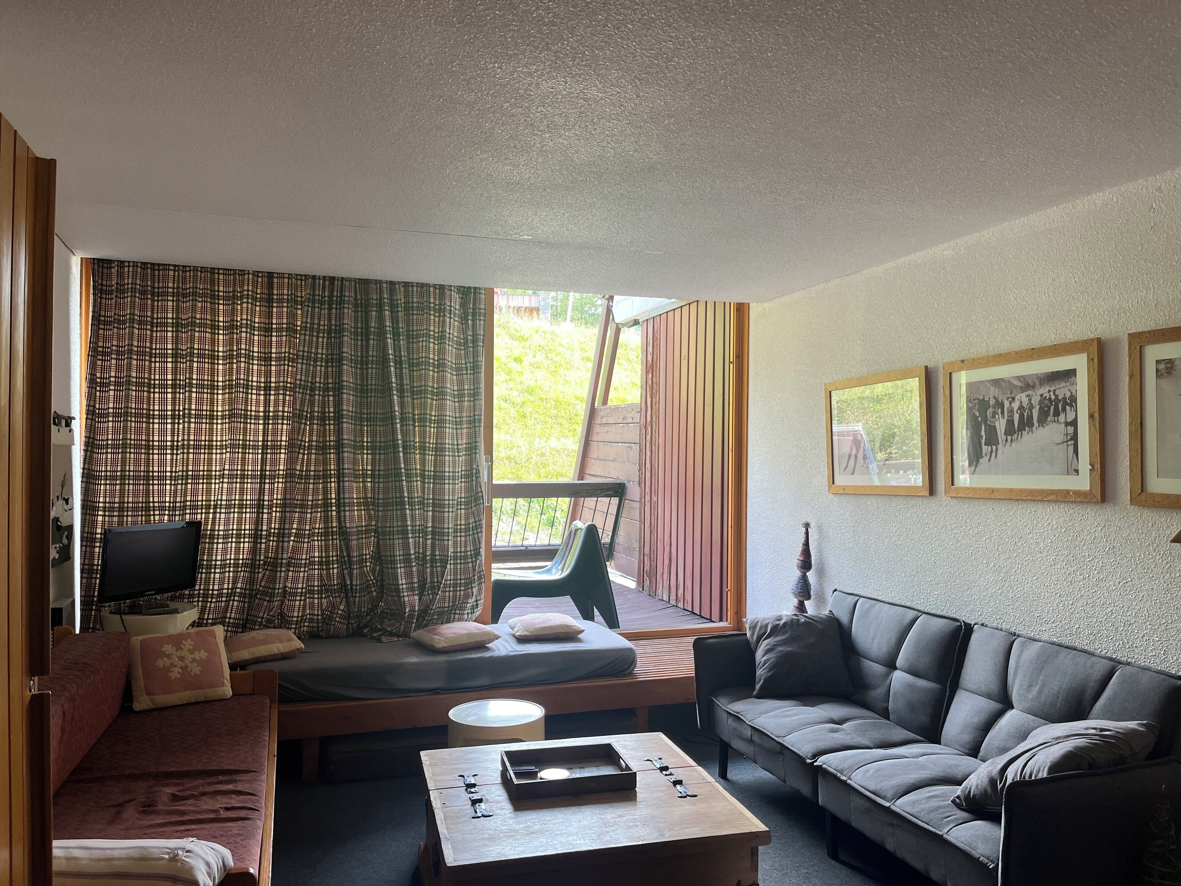 Ein gemütliches Zimmer mit einer Couch, einem kleinen Tisch, einem Fernseher und einem Fenster mit Vorhängen. An den Wänden hängen gerahmte Bilder.