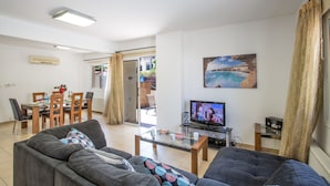 A living room with a sofa, a coffee table, a TV, and a dining area with chairs and a table. - Protaras Villa PV5 - Modern 3BDR Central Protaras Villa with Private Pool (Unknown)