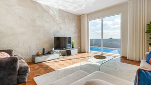 A modern living room with a flat-screen TV, a sofa, a glass coffee table, and a view of a swimming pool through large windows. - Funchal Bay View Villa by Madeira Sun Travel (Funchal)