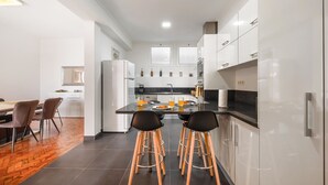 A modern kitchen with white cabinets, a central island with stools, and a dining area with a wooden table and chairs. - Funchal Bay View Villa by Madeira Sun Travel (Funchal)