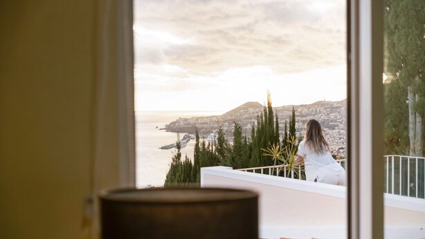 A balcony with a view of a coastal cityscape, a person sitting, and a potted plant. - Funchal Bay View Villa by Madeira Sun Travel (Funchal)
