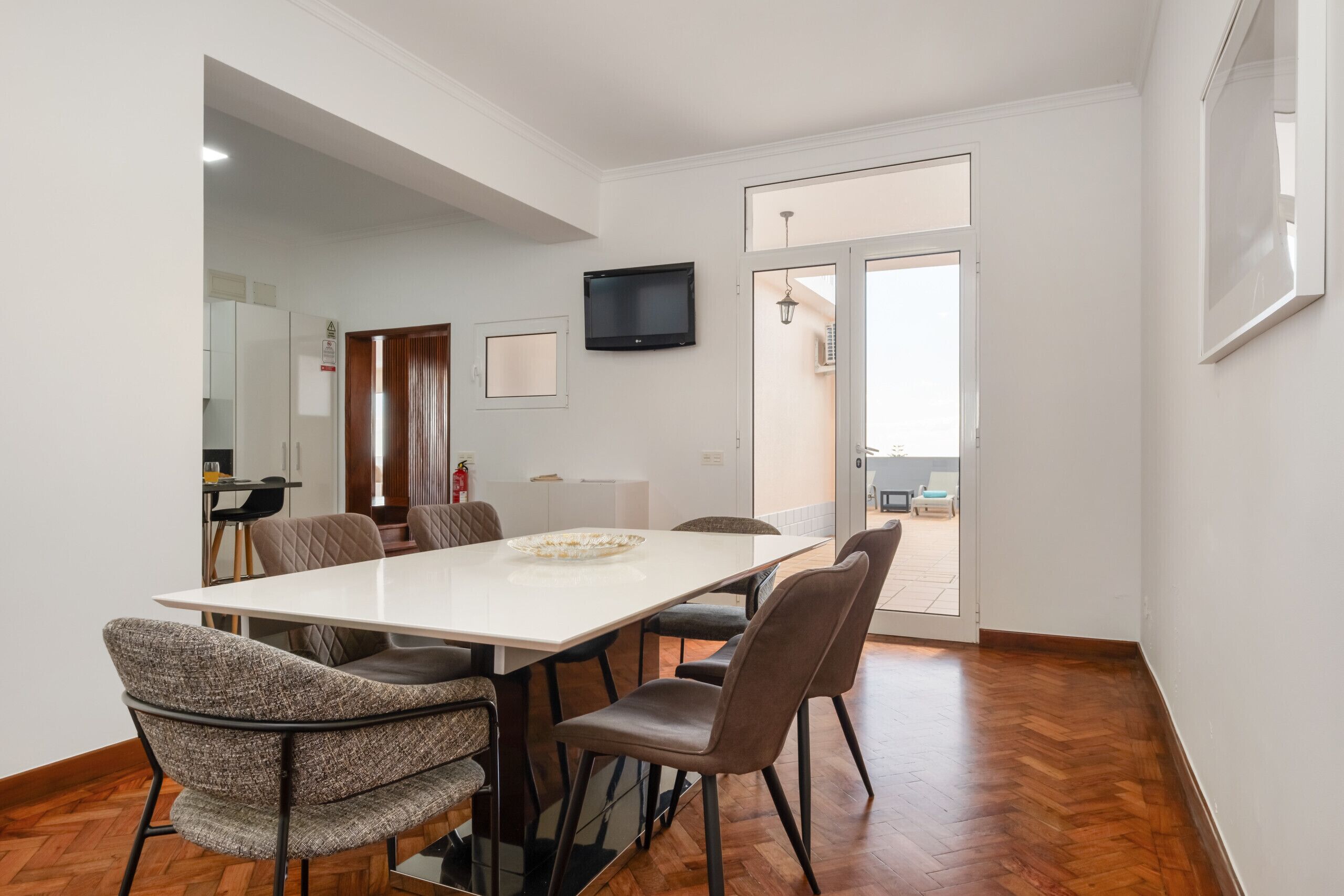 A dining area with a white table, six chairs, a television on the wall, and a door leading to an outdoor space.