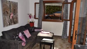 A living room with a dark brown sofa, a small table, and a window with a view of a car and greenery outside.