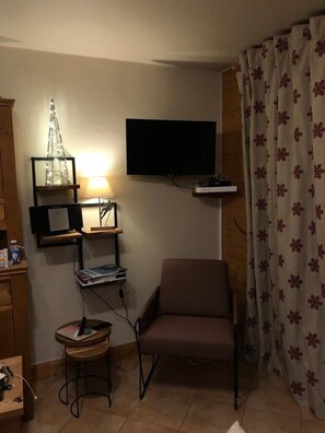 Una habitación pequeña y acogedora con un televisor montado en la pared, una silla, una mesita y una lámpara decorativa.