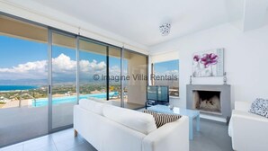 Un salón amplio con un sofá blanco, una mesa de centro y una chimenea. Un gran ventanal corredera da a un balcón con piscina y vistas al mar.