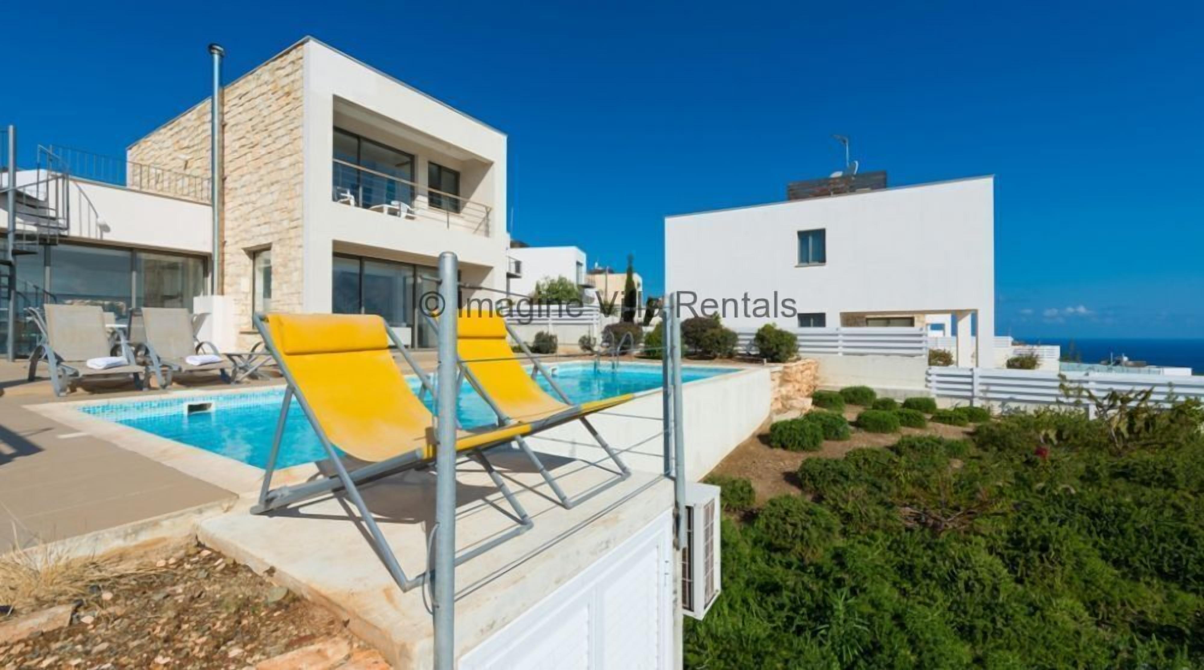 Un bâtiment moderne doté d’une piscine, de chaises longues jaunes et offrant une vue sur la mer.