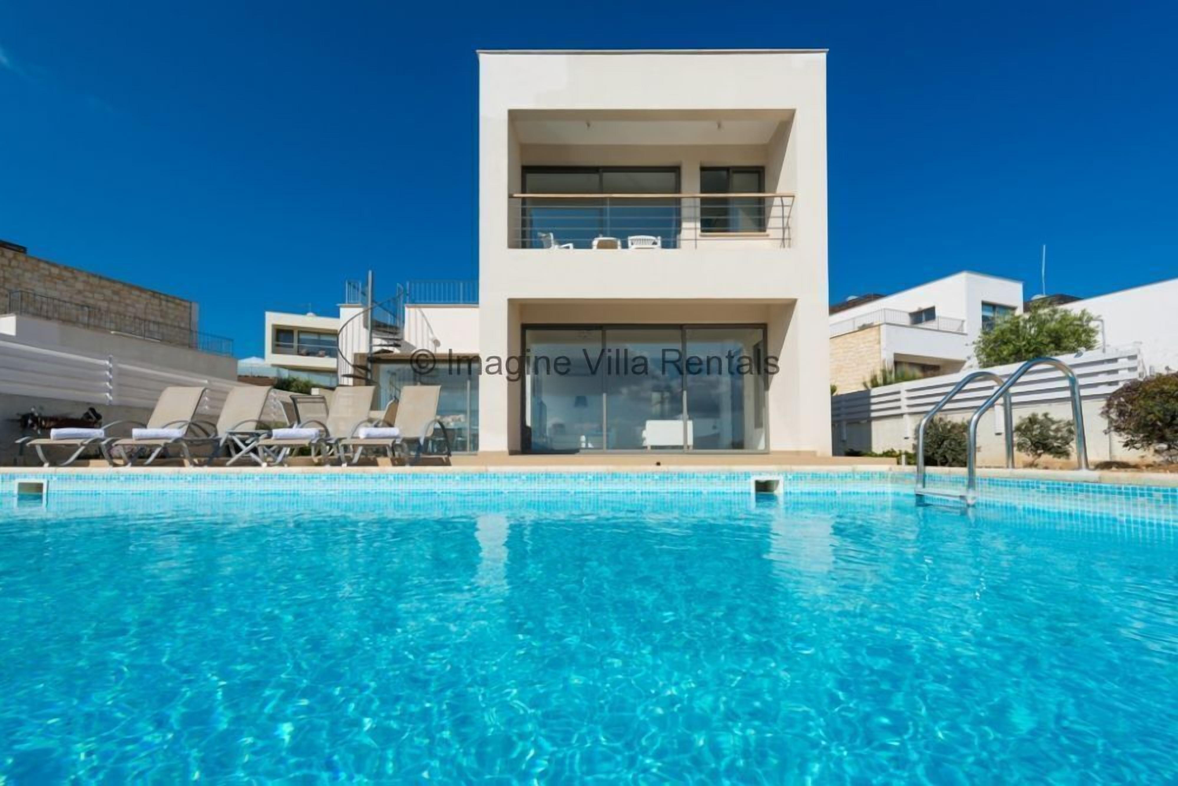 A modern villa with a swimming pool, a balcony with sun loungers, and a clear blue sky.