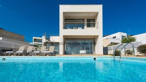 Una villa moderna con piscina, un balcón con tumbonas y un cielo azul despejado.