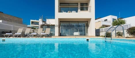 A modern villa with a swimming pool, a balcony with sun loungers, and a clear blue sky.