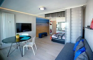 Une chambre d’hôtel moderne équipée d’une télévision à écran plat, d’une table à manger avec des chaises, d’un canapé, d’un lit avec une grande fenêtre et d’une petite plante en pot.
