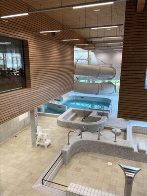 Un espace piscine intérieur moderne, doté de plusieurs éléments aquatiques, notamment une piscine centrale et des bains à remous tout autour, avec un lambris en bois et un revêtement de sol carrelé.