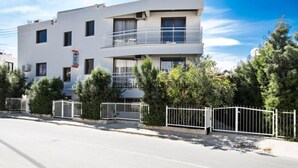 A modern two-story building with balconies, surrounded by a white fence and greenery.
