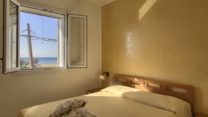 Quarto com cama, mesa de cabeceira, luminária na parede e janela com vista para a praia e uma cerca de arame farpado.