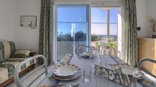 Uma área de refeições com uma mesa posta para quatro pessoas, um sofá e vista para o mar através de uma porta de vidro deslizante.