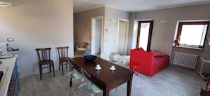 A dining area with a wooden table, chairs, and a blue bowl. There is a red sofa in the background and a window with a view.