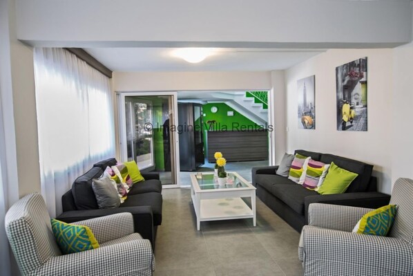 A modern living room with two black sofas, a glass coffee table, and checkered armchairs. A sliding glass door leads to a green area with a staircase.