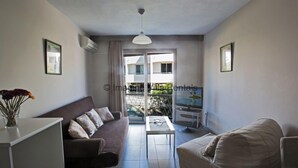 Un salon moderne avec un canapé, une table basse et un fauteuil poire. Une porte-fenêtre coulissante donne accès à un balcon d’où l’on peut admirer les bâtiments et la verdure.