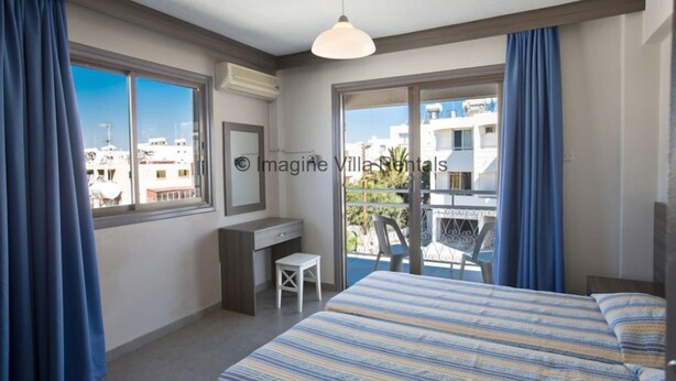 Een slaapkamer met een bed, een bureau en een stoel. Er is een balkon met uitzicht op gebouwen.